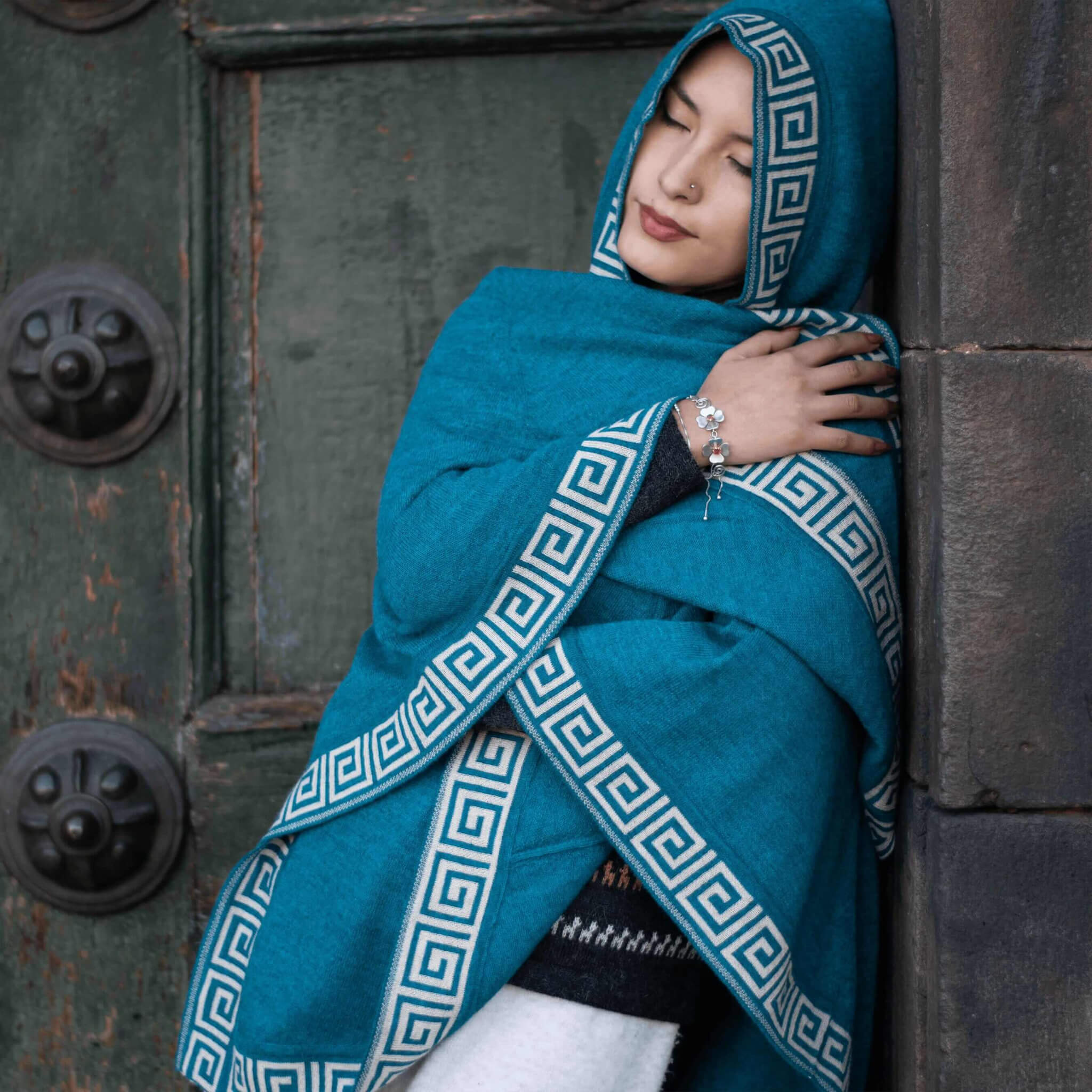 Weibliches Modell hüllt sich in den Alpaka Poncho mit Kapuze Luz in dunkeltürkis, gefertigt in der kleinen Alpakin Manufaktur in Peru