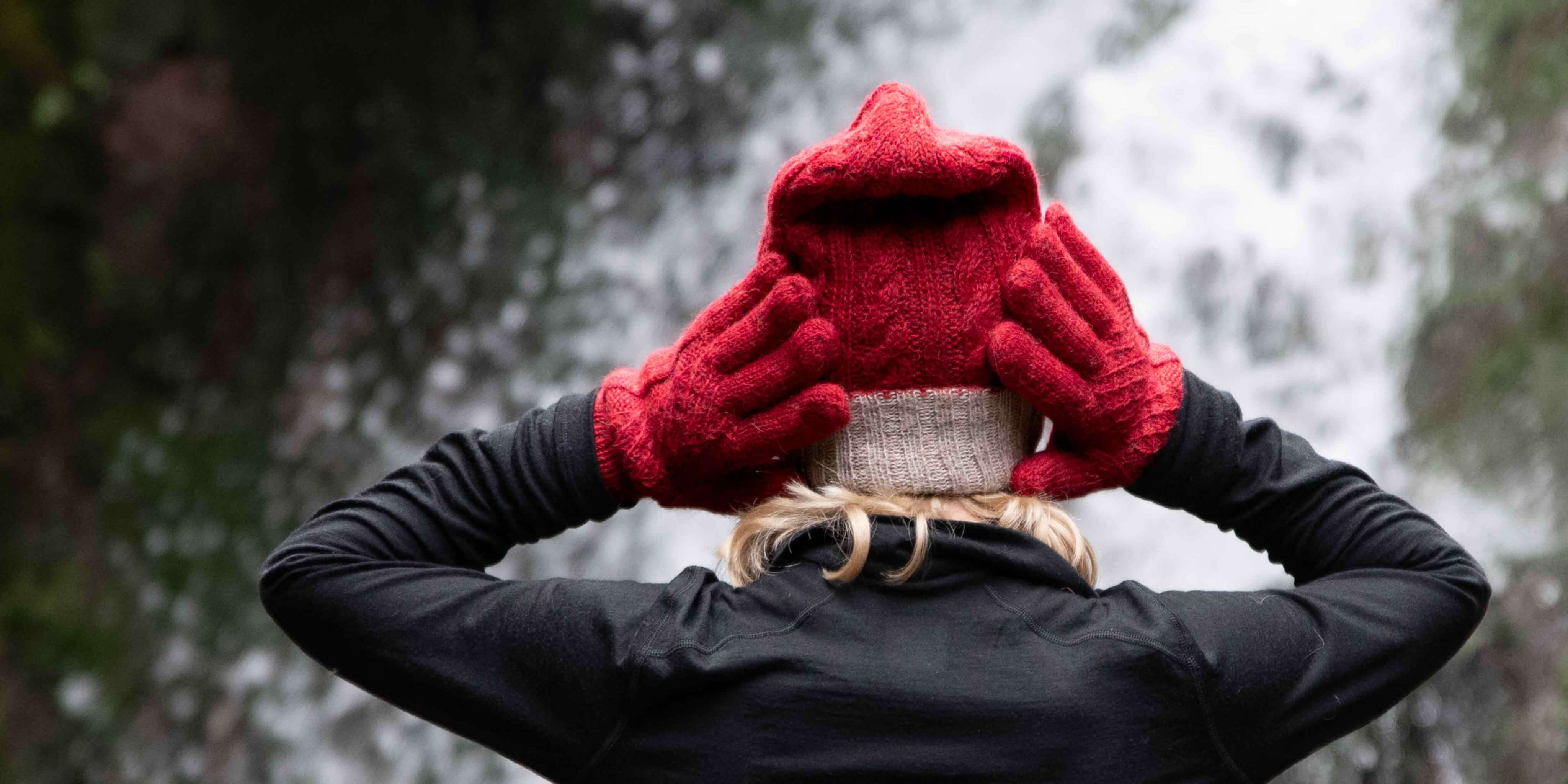 Model vor Wasserfall Kulisse fasst sich mit ihren Alpaka Wendehandschuhen von Alpakin in rot an ihre Mütze