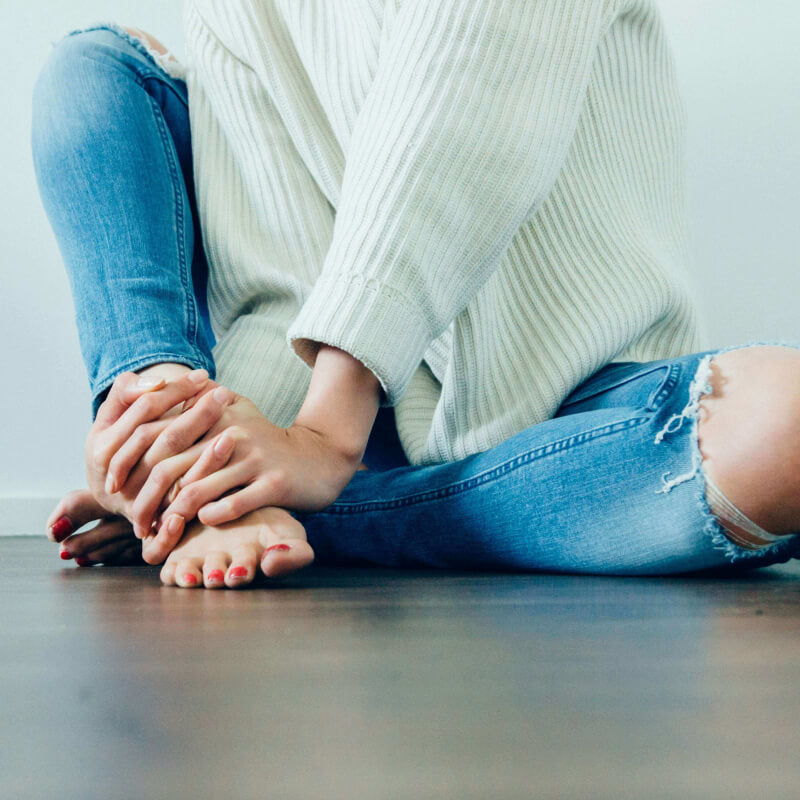 Frau mit Jeans und Pulli sitzt gemütlich auf dem Boden und legt die Hände auf die Füße