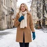 Frau trägt dunkeltürkis-farbige Alpaka Handschuhe mit Mantel und cremefarbigen Schal vor Stadtkulisse im Winter