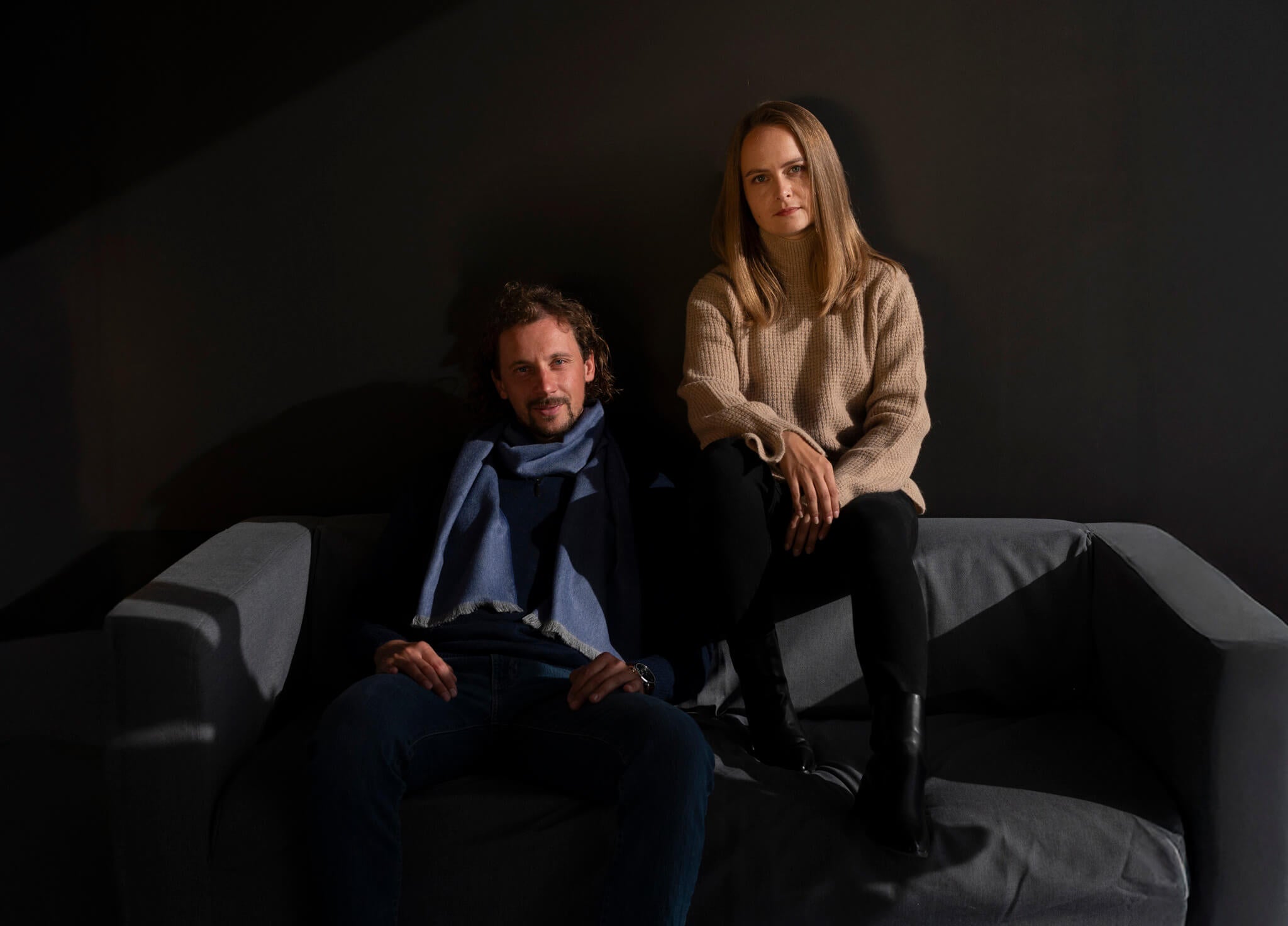 Mann und Frau sitzen gemeinsam auf einem schwarzen Sofa und tragen elegante Kleidung aus Alpakawolle von Alpakin