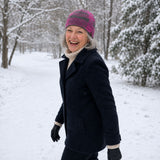 Frau mit grauen Haaren trägt in verschneiter Winterlandschaft ihre warme Merino Mütze in pink und lacht