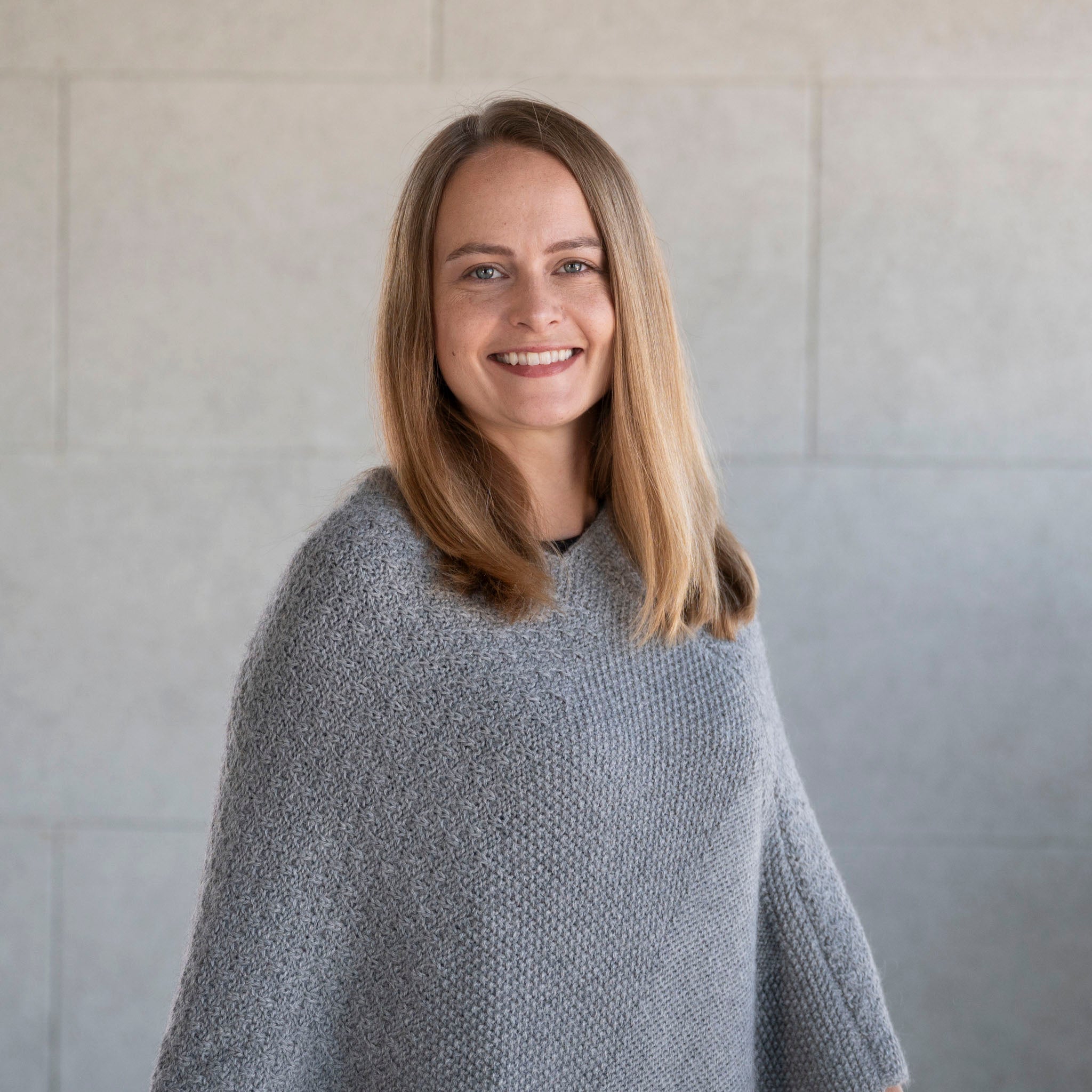 Blonde Frau trägt den weichen Naira Alpaka Strickponcho in grau mit feiner Musterung im Textil