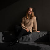 Frau auf schwarzer Couch trägt den Nuna Alpaka Rollkragenpullover von Alpakin in Naturfarbe hellbraun, ohne chemische Färbung